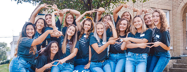 A group of smiling young women standing close together outdoors, wearing matching dark t-shirts and blue jeans, making hand signs and posing cheerfully in front of a brick building and trees.