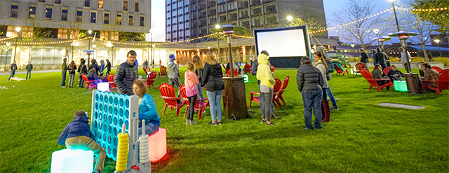 People gather on a grassy lawn in an urban area, playing giant games like Connect Four, sitting in red chairs, and watching an outdoor movie screen at dusk, surrounded by buildings and string lights.