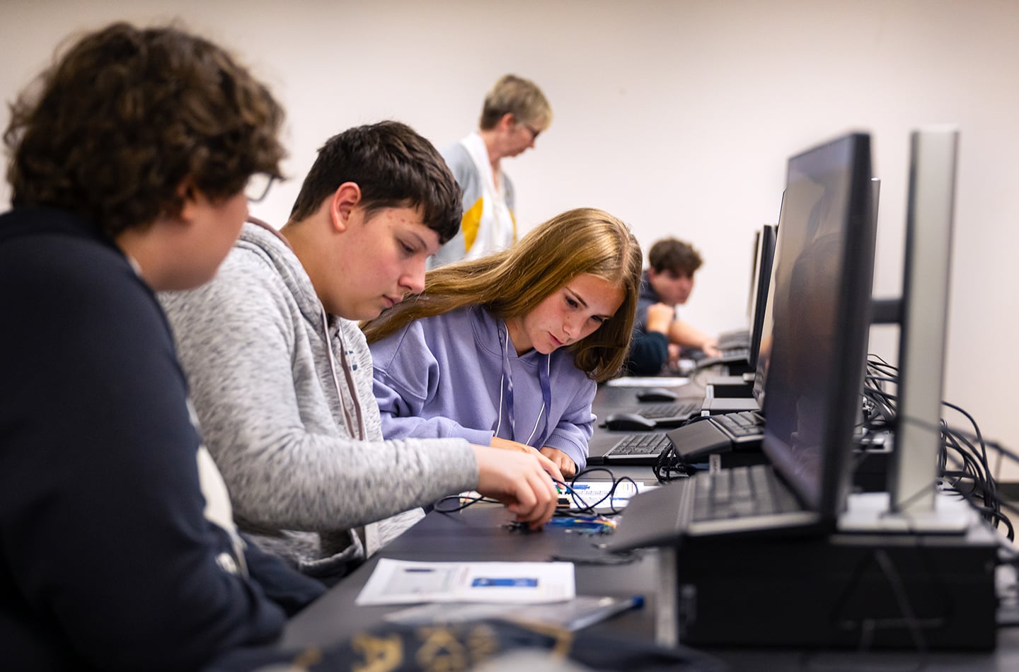 Three students in a computer lab working collaboratively on a hardware project. One student adjusts small electronic components and wiring on a desk while two others look on closely, with several computer monitors and an instructor visible in the background.
