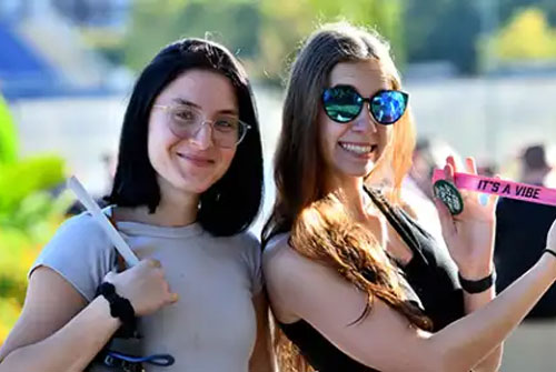 Two smiling college students pose outdoors on a sunny day, one holding up a pink wristband that reads 'It's a Vibe,' at a campus event.