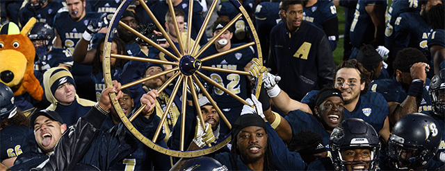 A jubilant college football team in navy uniforms celebrates on the field, holding up a large wooden wagon wheel trophy. A mascot in an animal costume is visible on the left, and everyone appears excited and joyful.