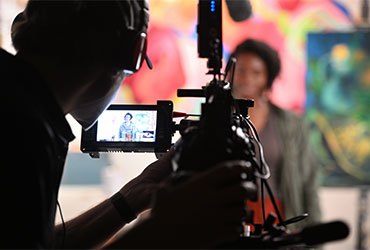 Producer films an interview with a camera while another speaks on camera in a media production setting.