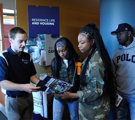 A staff member explains a brochure about residence life and housing to a student and family, two women and one man, standing indoors near an informational display. The students listen and look at the booklet.