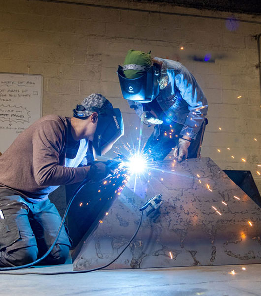 A Myers sculpture student works with artist to weld sections of a metal public sculpture in the studio.
