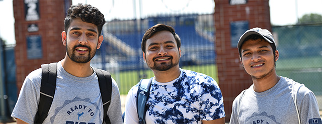 Three young men are smiling outdoors on a sunny day, wearing casual t-shirts and backpacks. A sports field and stadium seating are visible in the background behind a fence and brick pillars.