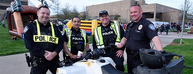 Four smiling police officers in uniform stand outdoors near an all-terrain vehicle, with inflatable attractions and buildings visible in the background at what appears to be a public event.