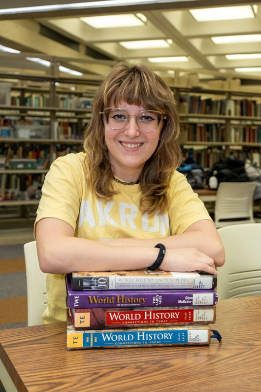 Student wearing an Akron shirt sits at a library table with a stack of world history textbooks.