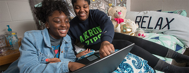 Two smiling students relax on a bed in a dorm room, looking at a laptop together. Behind them are a large pillow with RELAX written on it and a stuffed teddy bear. The room has cozy, decorated bedding.