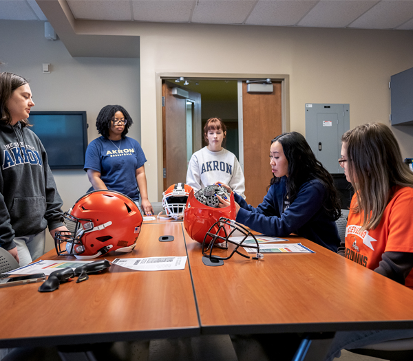 Five students gather around a table with football helmets and papers, discussing something. Some wear Akron shirts. The setting appears to be a classroom or meeting room with neutral walls and fluorescent lighting.