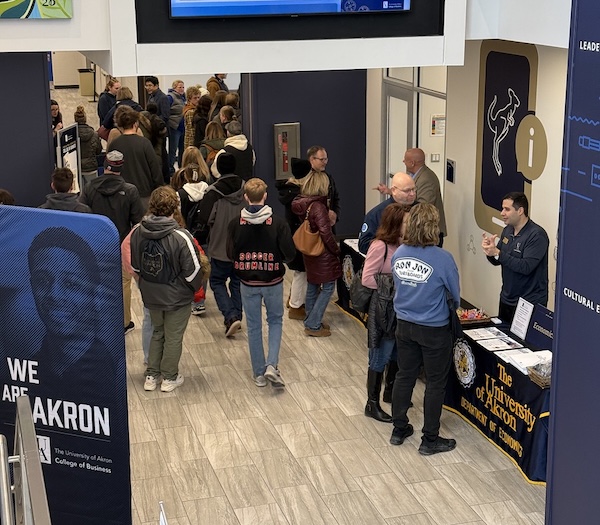 A group of people walk through a hallway at a university event, with informational tables set up along the side and staff members talking to attendees. Signs for The University of Akron and Iron Jay are visible.