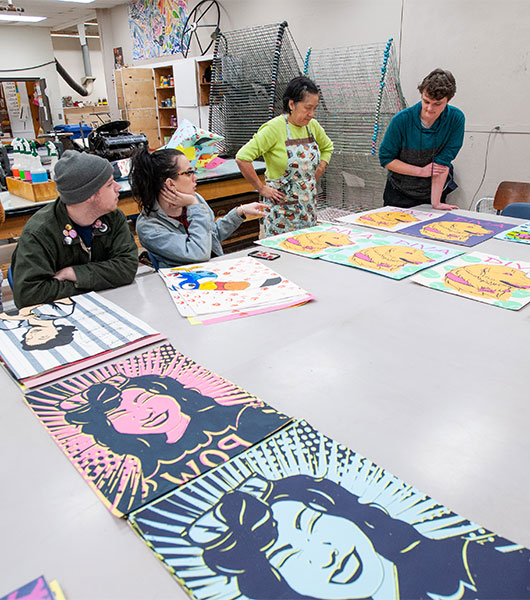 Printmaking students gather around a table displaying colorful prints and works in progress.