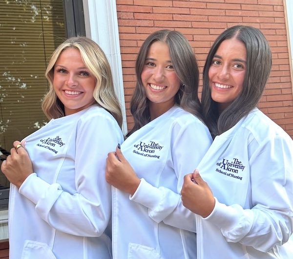 Three young women stand side-by-side, smiling and posing in white zip-up jackets. Each jacket features the embroidered logo for 