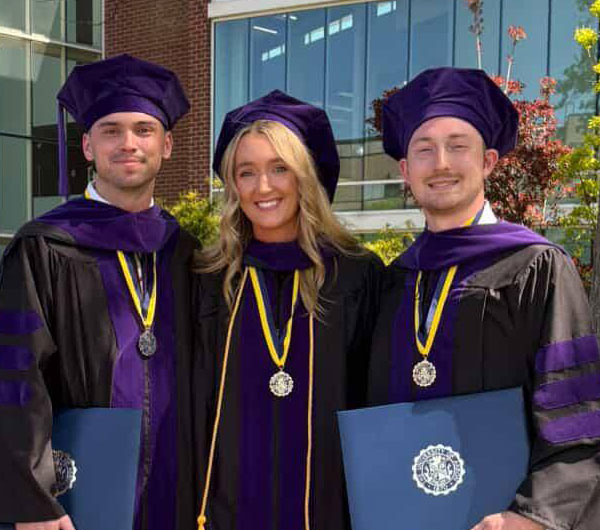 Three graduates wearing purple academic regalia and medals pose outdoors, one holding a diploma folder.