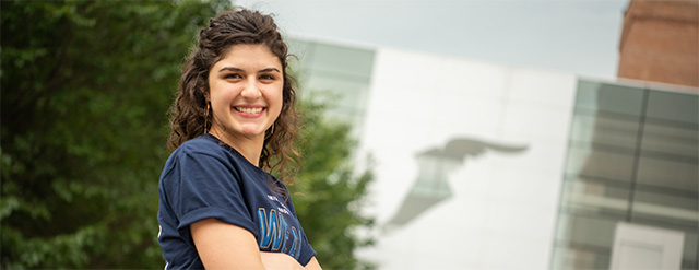 A young woman with curly dark hair smiles at the camera, standing outdoors in front of a modern building with a large wing-shaped shadow on the wall. Trees are visible on the left side.