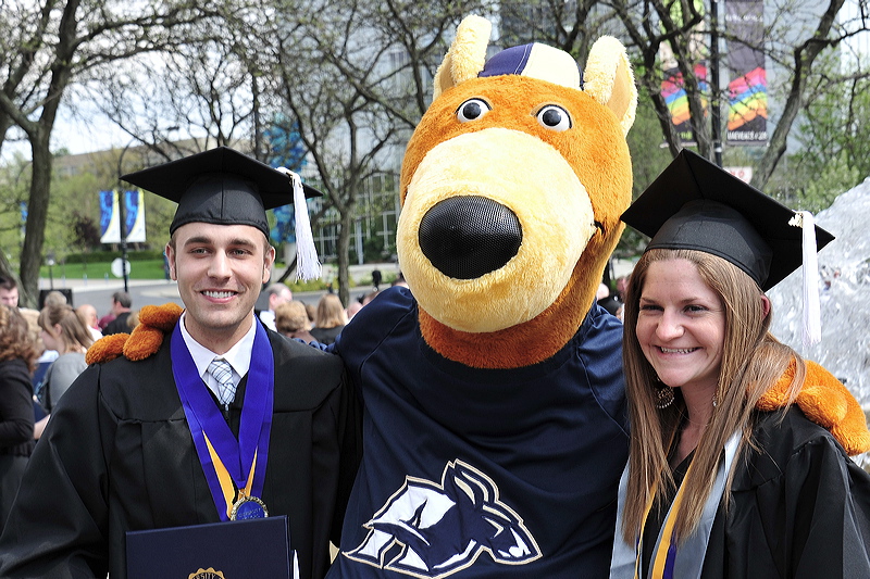 Two UA students in graduation gowns, standing with the University of Akron mascot - Zippy